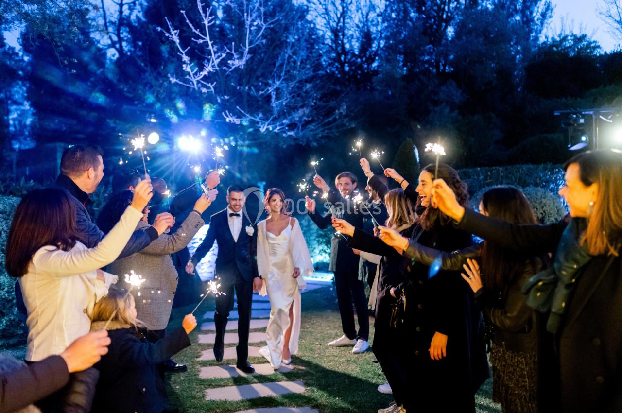 Un couple de mariés marche sur une allée extérieure, entouré d'invités tenant des cierges magiques allumés.