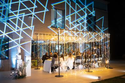 Table dressée pour un dîner en extérieur sous une structure lumineuse avec guirlandes, près d'un bâtiment moderne.