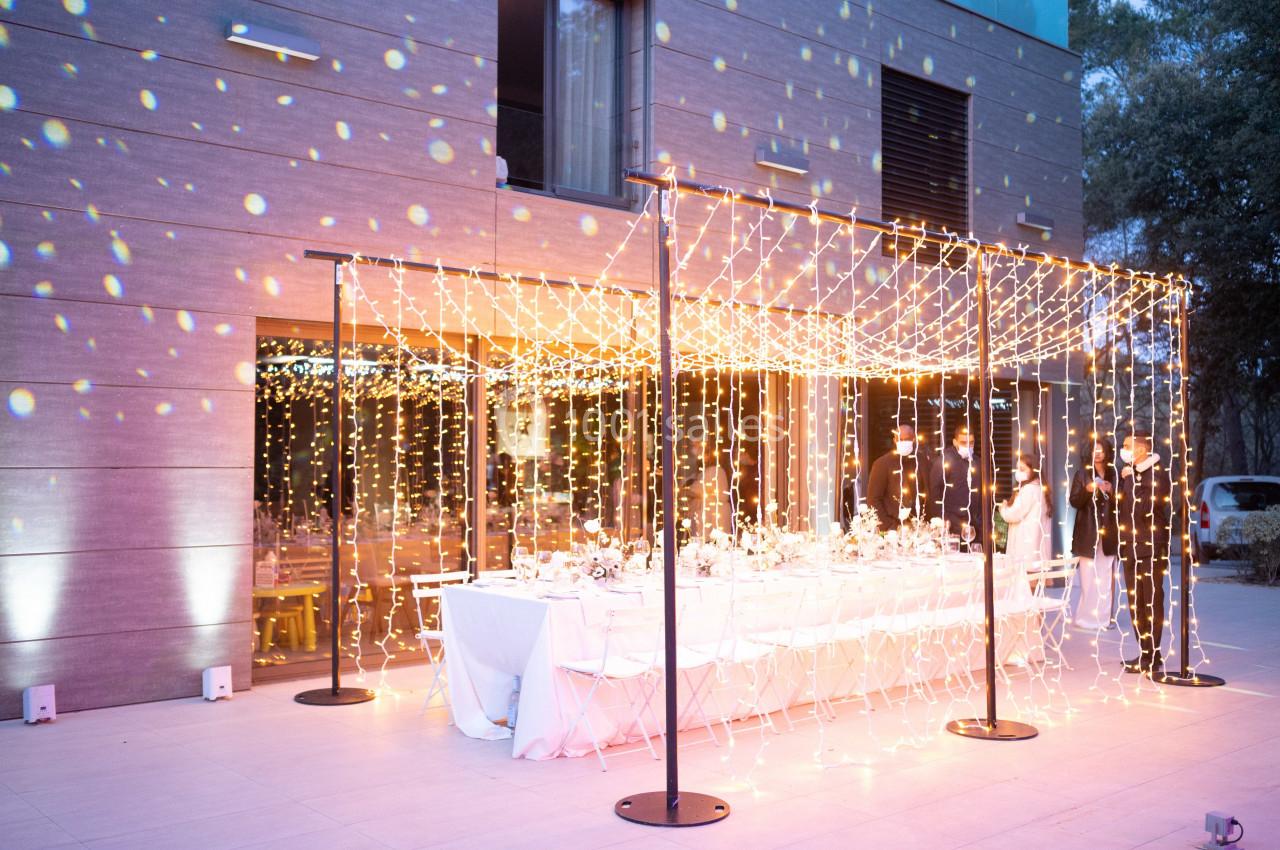 Table dressée pour un dîner en extérieur sous une structure lumineuse avec guirlandes, près d'un bâtiment moderne.
