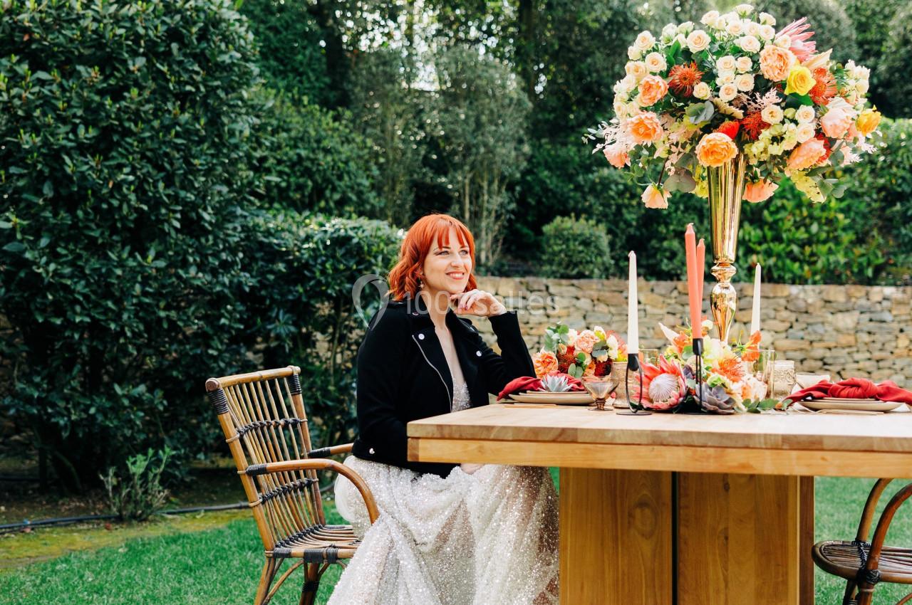Une femme en robe blanche assise à une table décorée de fleurs colorées dans un jardin verdoyant.