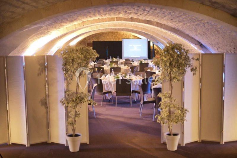 Salle voûtée aménagée pour un événement, avec des tables rondes dressées et un écran de projection en fond.