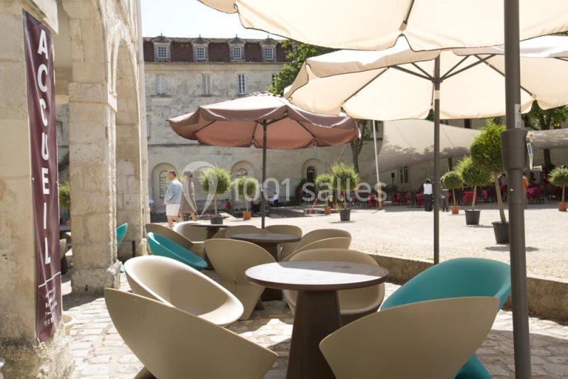 Terrasse extérieure avec tables, chaises design et parasols, située devant un bâtiment en pierre avec une bannière ’Accueil’.