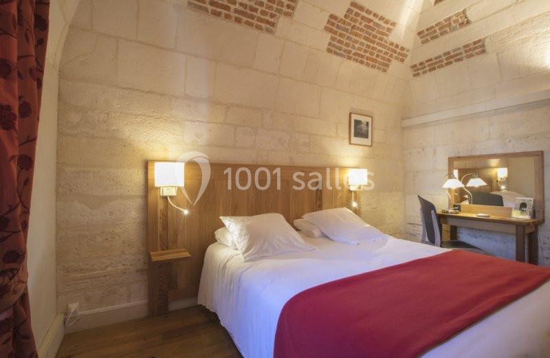 Chambre avec lit double, drap rouge, murs en pierre, bureau en bois et éclairage doux.