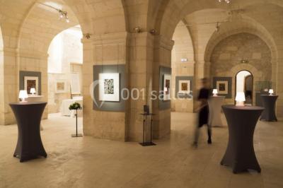 Deux lits simples avec draps rouges dans une chambre lumineuse en pierre, équipée d'une fenêtre et d'un bureau.