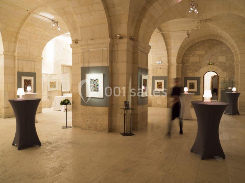 Salle voûtée en pierre avec des œuvres d'art accrochées aux murs et des tables hautes éclairées.
