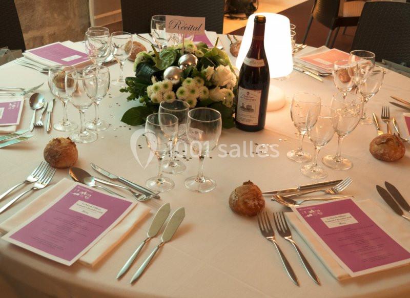 Table dressée avec nappes blanches, menus, verres, couverts, centre de table floral, bouteille de vin et petits pains.