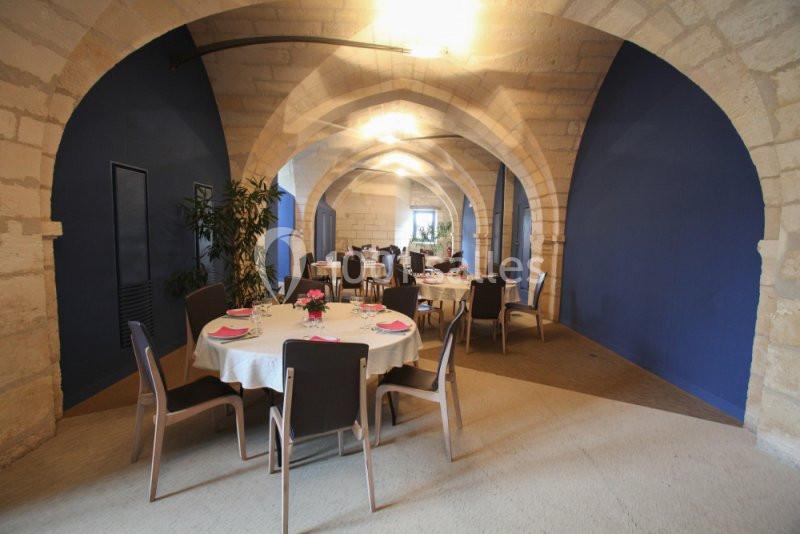 Salle voûtée en pierre avec tables rondes dressées, chaises modernes et murs peints en bleu.
