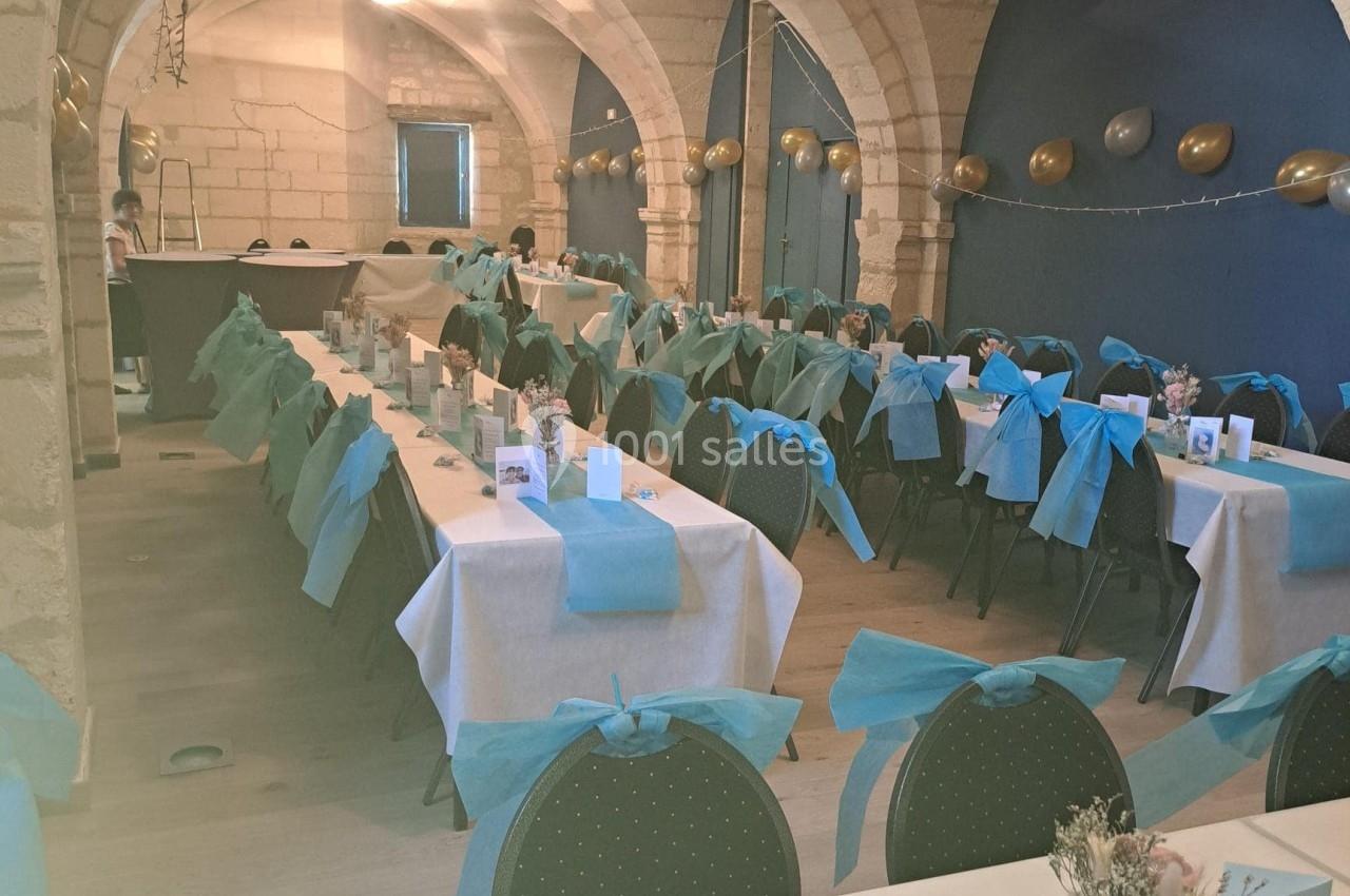 Salle voûtée décorée pour un événement, avec des tables alignées, nappes blanches et bleues, et chaises assorties.