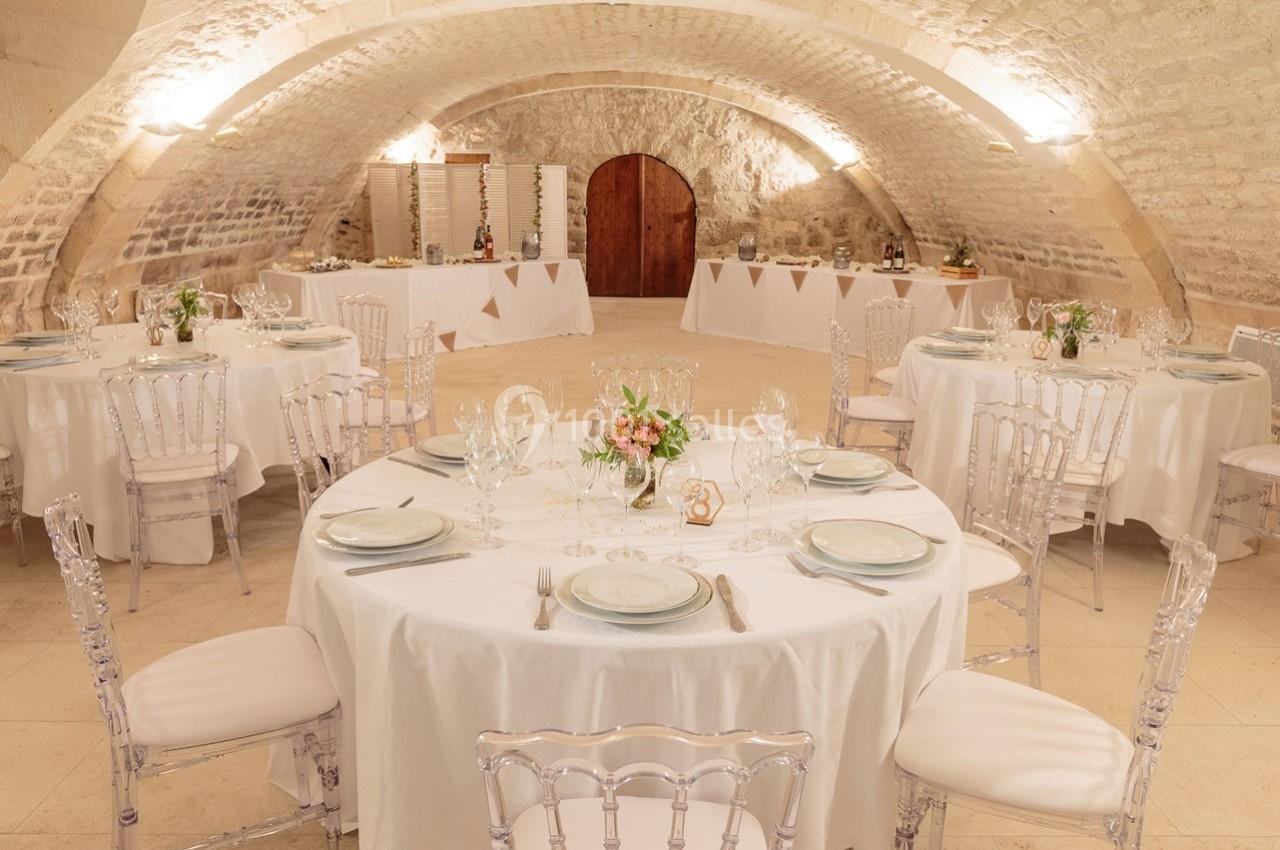 Salle voûtée en pierre avec tables rondes dressées pour un repas, décorations florales et lumière chaleureuse.