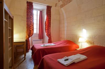 Deux lits simples avec draps rouges dans une chambre lumineuse en pierre, équipée d'une fenêtre et d'un bureau.