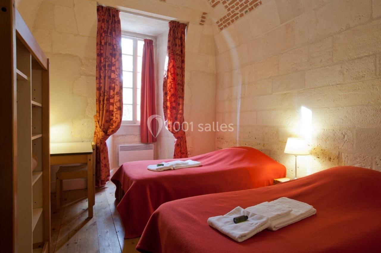 Deux lits simples avec draps rouges dans une chambre lumineuse en pierre, équipée d'une fenêtre et d'un bureau.