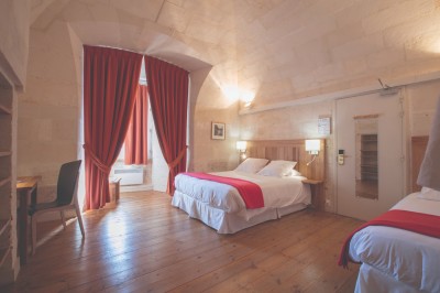 Deux lits simples avec draps rouges dans une chambre lumineuse en pierre, équipée d'une fenêtre et d'un bureau.