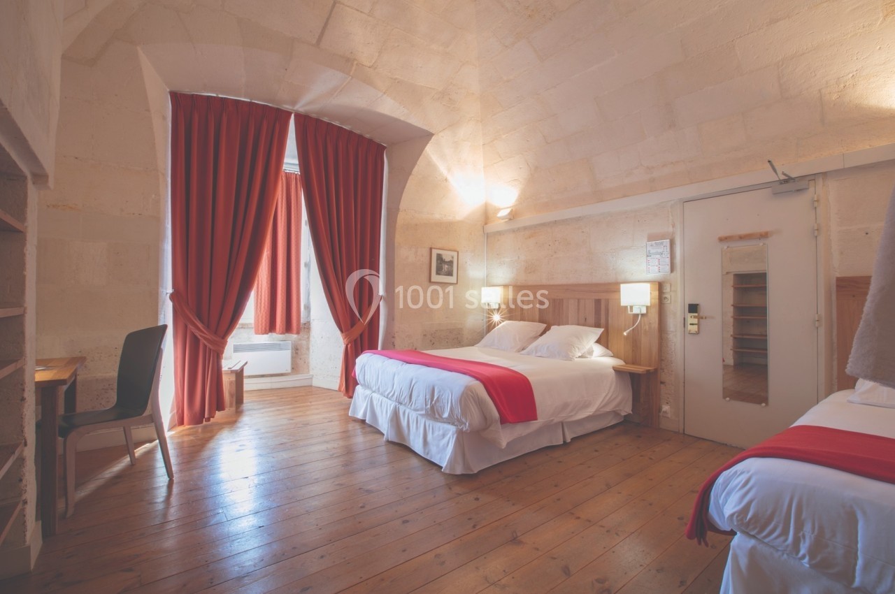 Chambre lumineuse avec murs en pierre, deux lits, rideaux rouges et parquet en bois clair.