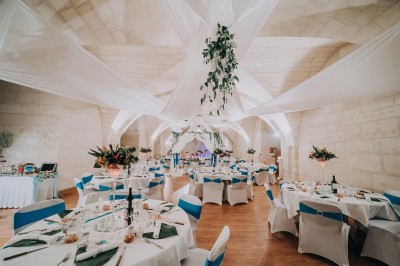 Deux lits simples avec draps rouges dans une chambre lumineuse en pierre, équipée d'une fenêtre et d'un bureau.