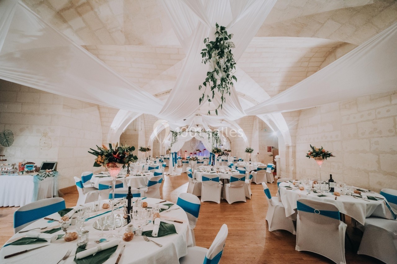 Salle de réception décorée avec des tables dressées, des nappes blanches, des chaises bleues et des drapés suspendus.