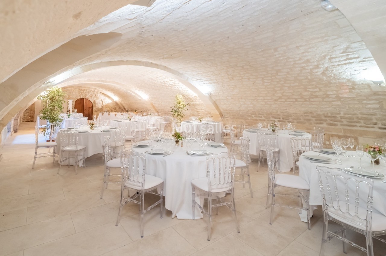 Salle voûtée en pierre avec tables rondes dressées pour un événement, décorée de fleurs et éclairée par des appliques…