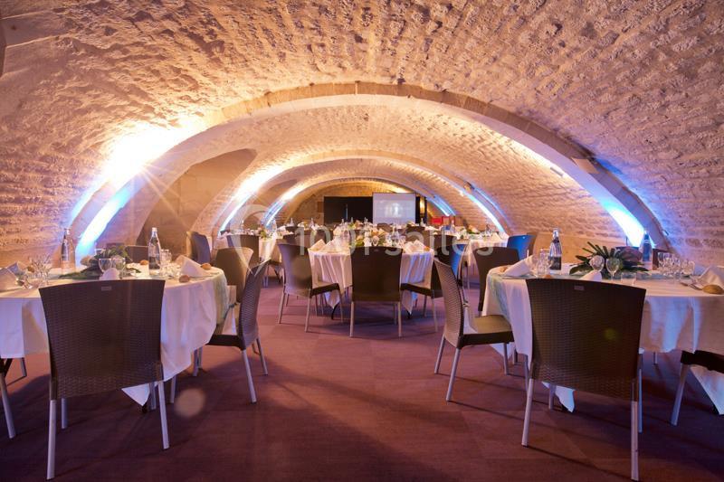 Salle voûtée éclairée avec des tables rondes dressées pour un événement.