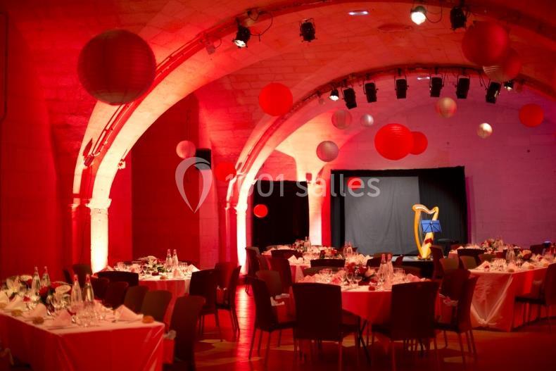 Salle voûtée éclairée en rouge, tables rondes dressées pour un événement, harpe et décorations suspendues.
