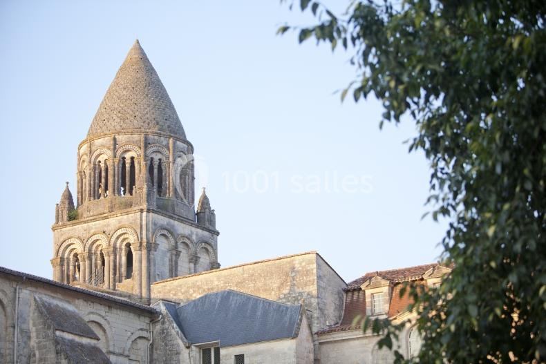 Clocher en pierre d'une église romane, entouré de bâtiments anciens et d'arbres sous un ciel clair.