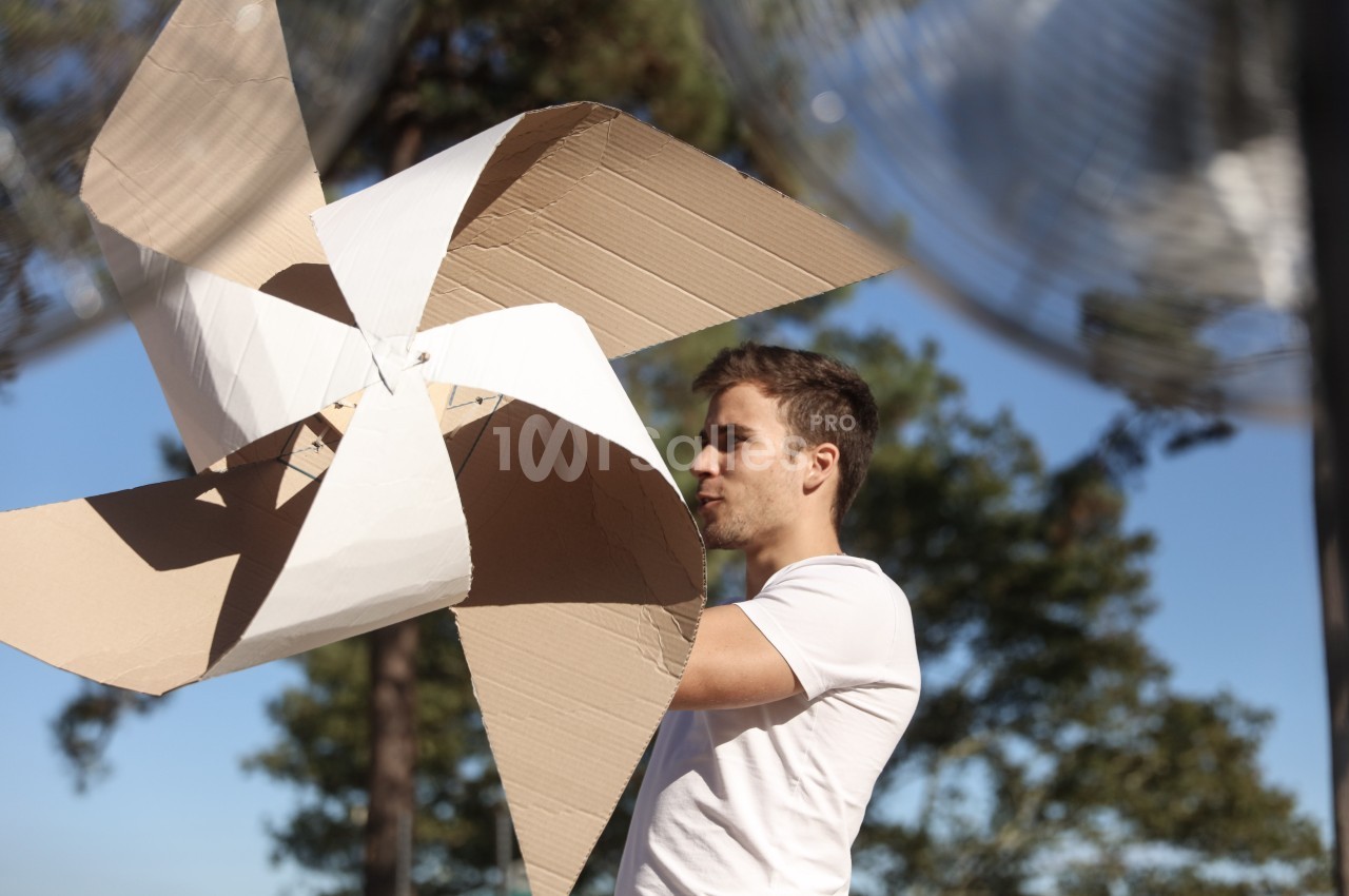 Un homme en t-shirt blanc tient une grande éolienne en carton dans un environnement extérieur ensoleillé.