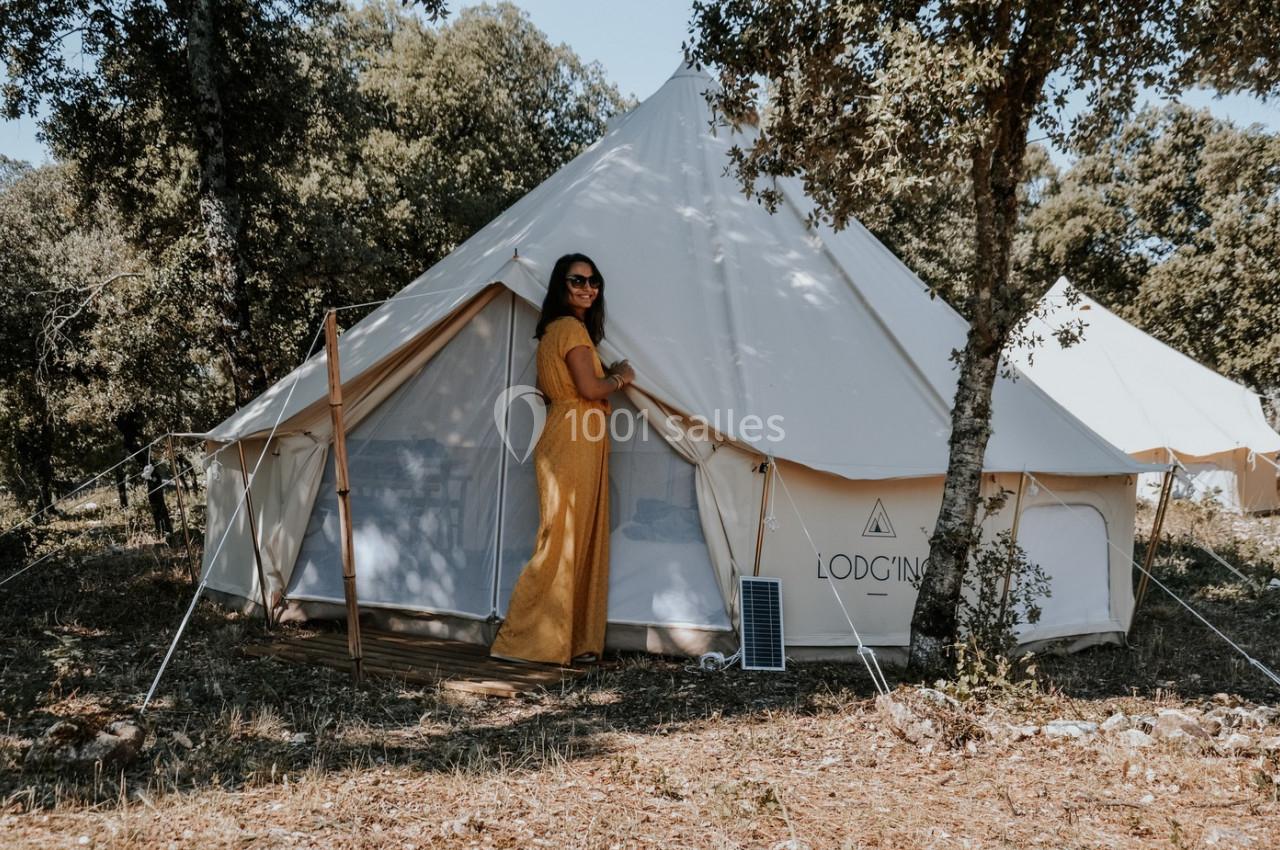Femme en robe jaune devant une tente blanche installée dans un cadre naturel boisé.