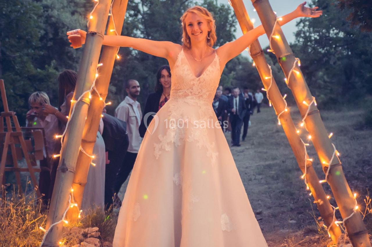 Une femme en robe de mariée sourit, bras ouverts, sous une arche décorée de lumières dans un cadre extérieur boisé.