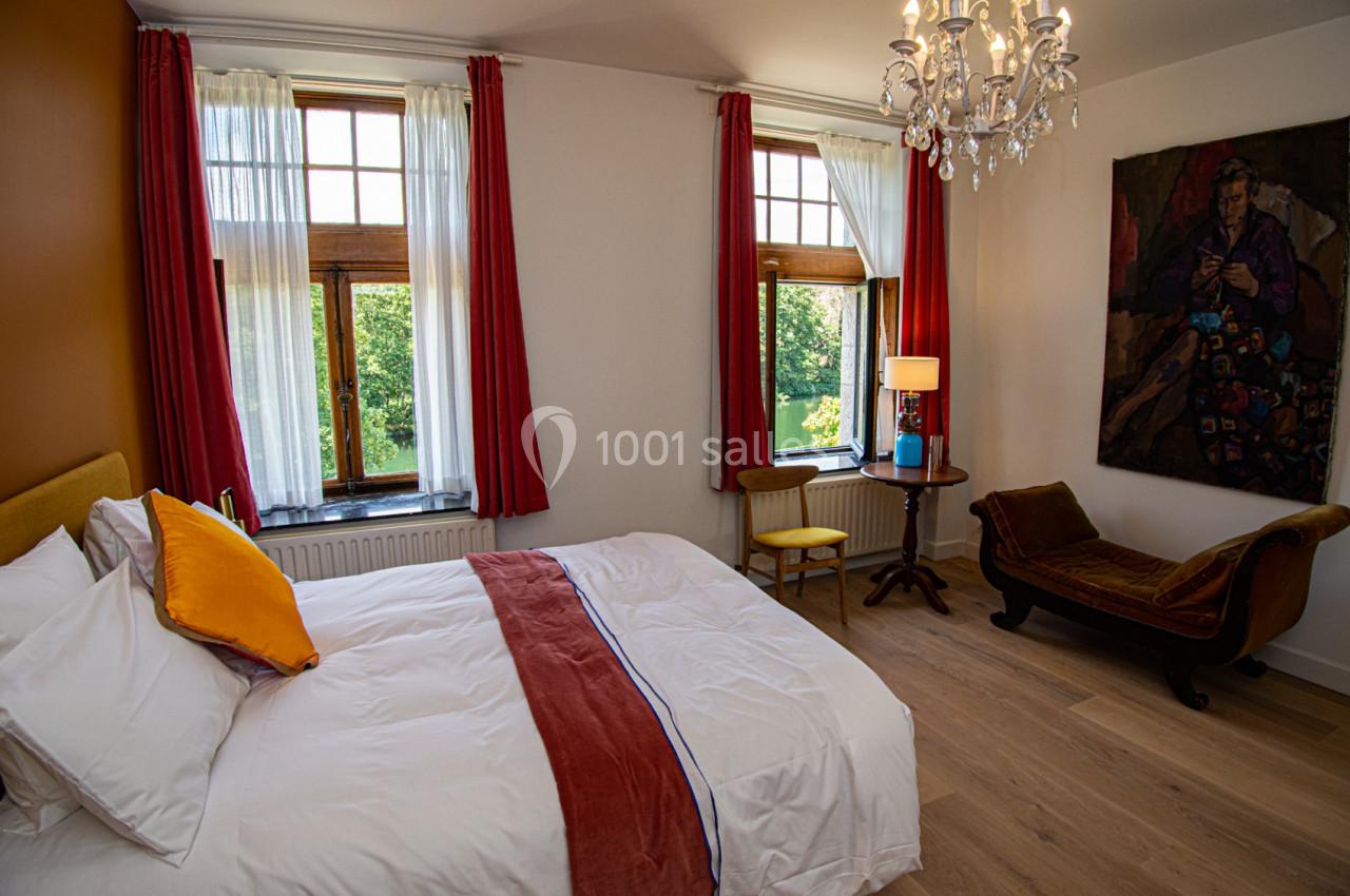 Chambre lumineuse avec lit double, fauteuil, table avec lampe, grandes fenêtres et vue sur un jardin verdoyant.