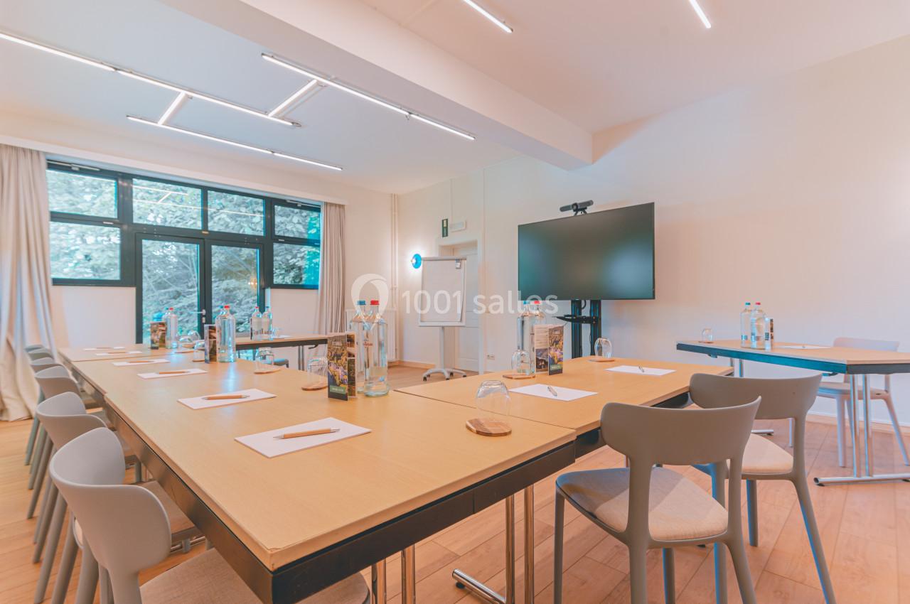 Salle de réunion lumineuse avec tables en bois disposées en U, chaises grises, écran, paperboard et bouteilles d'eau.