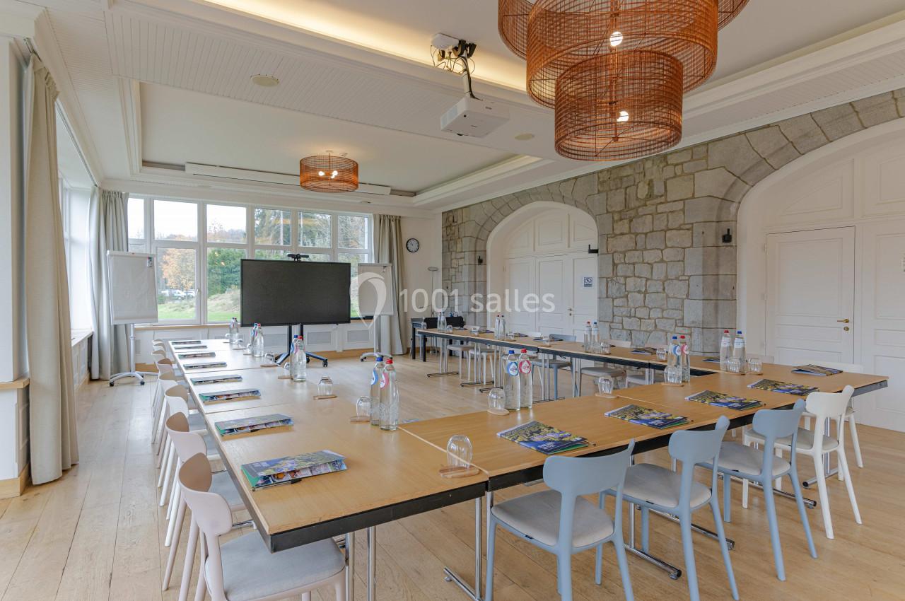 Salle de réunion lumineuse avec grandes fenêtres, tables en U, chaises colorées et écran de présentation.