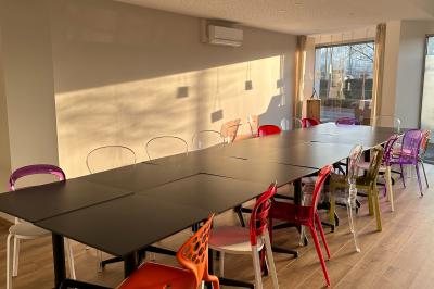 Salle lumineuse avec de grandes tables noires, chaises colorées et parquet en bois, éclairée par la lumière du soleil.