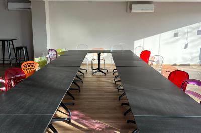 Salle lumineuse avec de grandes tables noires, chaises colorées et parquet en bois, éclairée par la lumière du soleil.