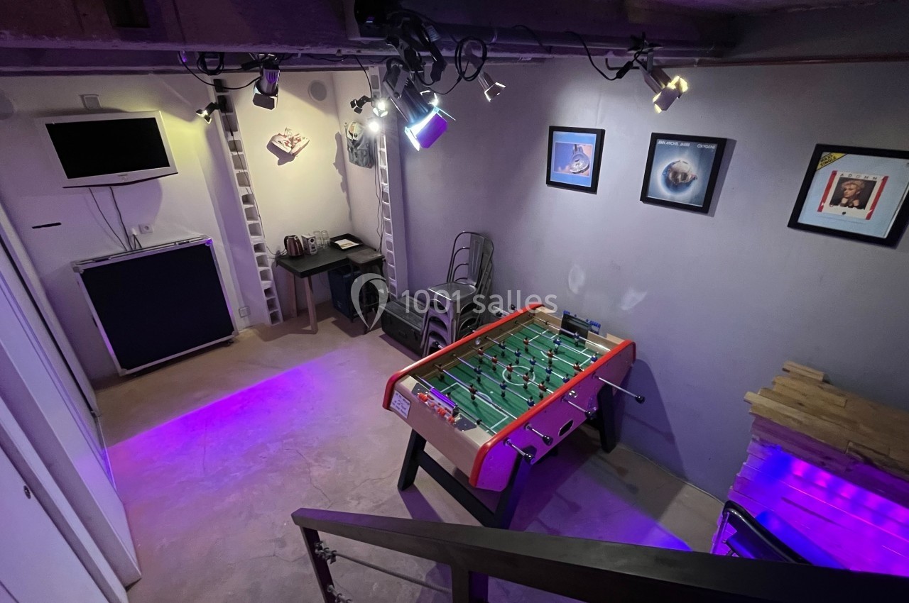 Salle de loisirs avec baby-foot, éclairage violet, télé murale, décorations et coin café.