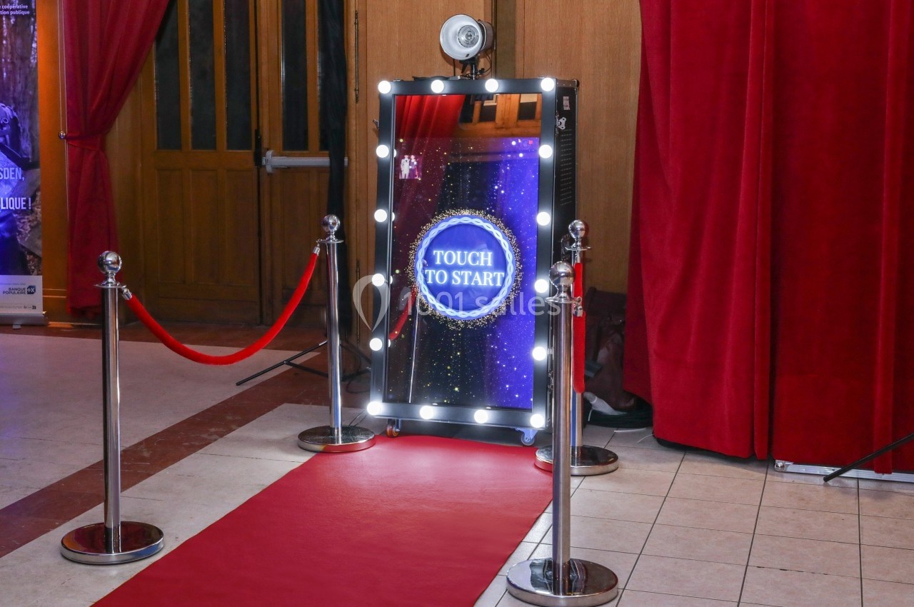 Miroir interactif lumineux avec écran tactile, entouré de rideaux rouges et d'un tapis rouge dans un espace intérieur.