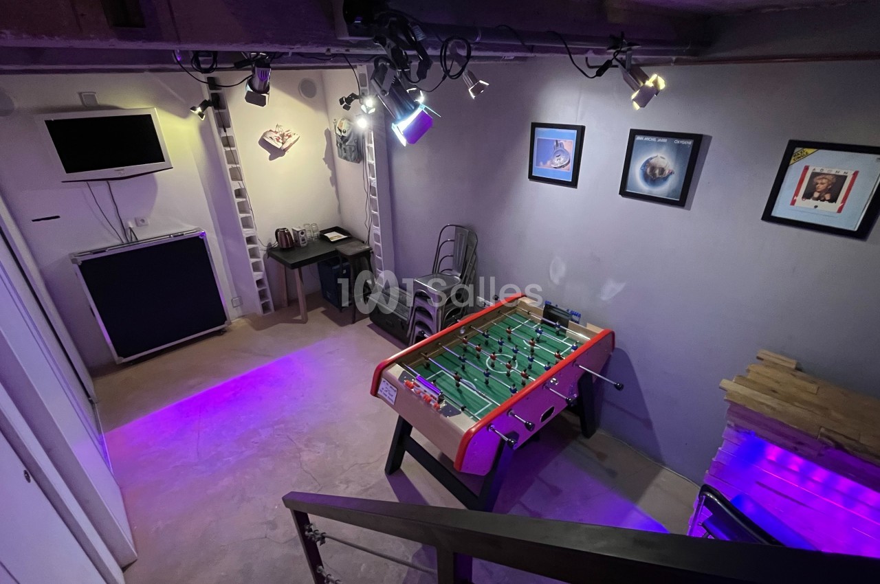 Salle de loisirs avec baby-foot, éclairage violet, télé murale, décorations et coin café.