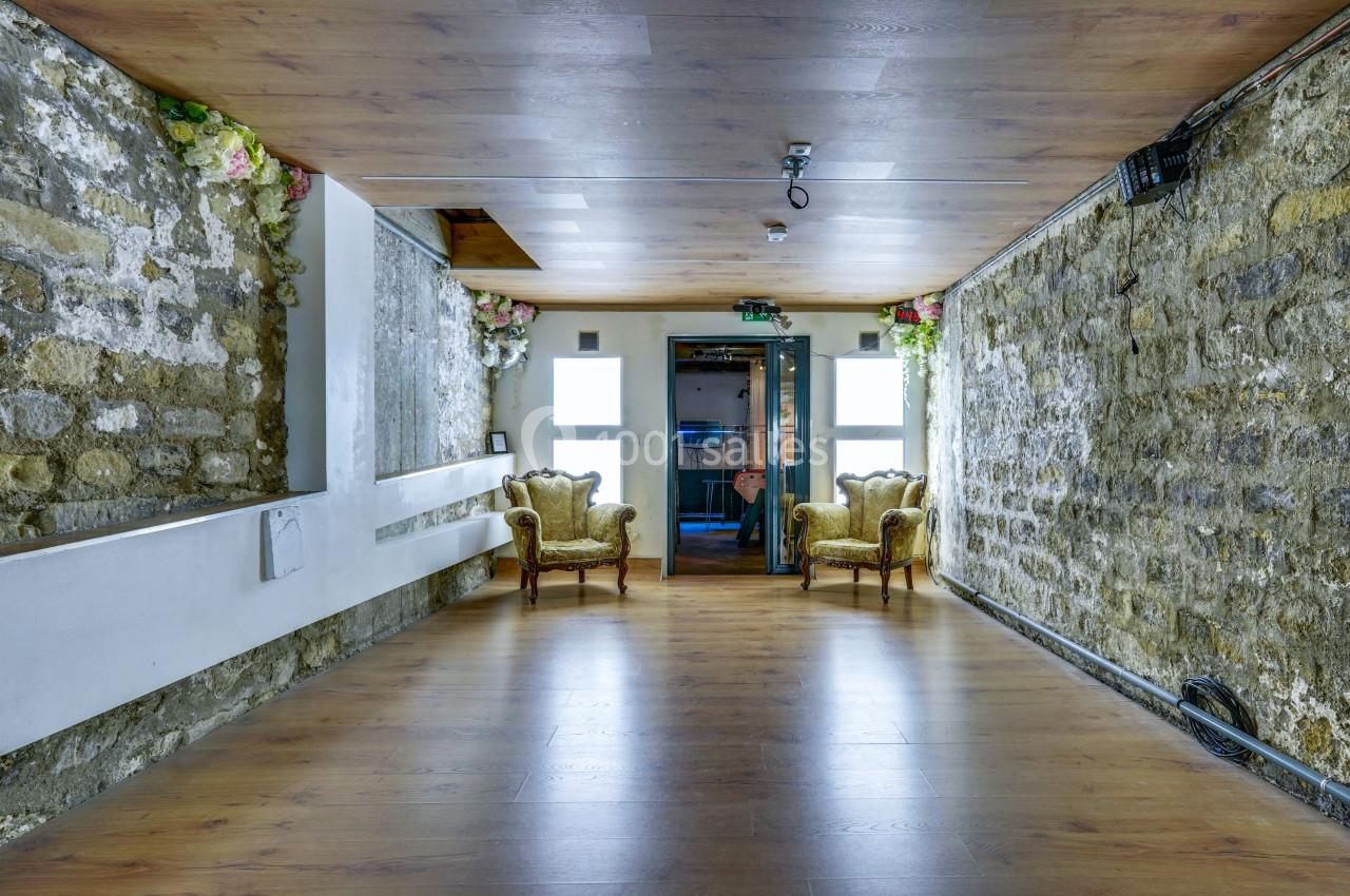 Salle vide avec murs en pierre, parquet au sol, plafond en bois et deux fauteuils anciens près d'une porte vitrée.