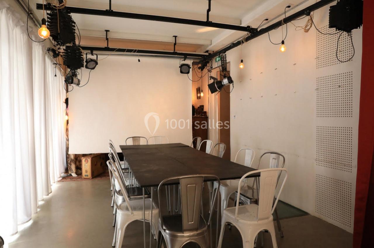 Salle de réunion avec table rectangulaire, chaises métalliques, éclairage suspendu et écran blanc en arrière-plan.