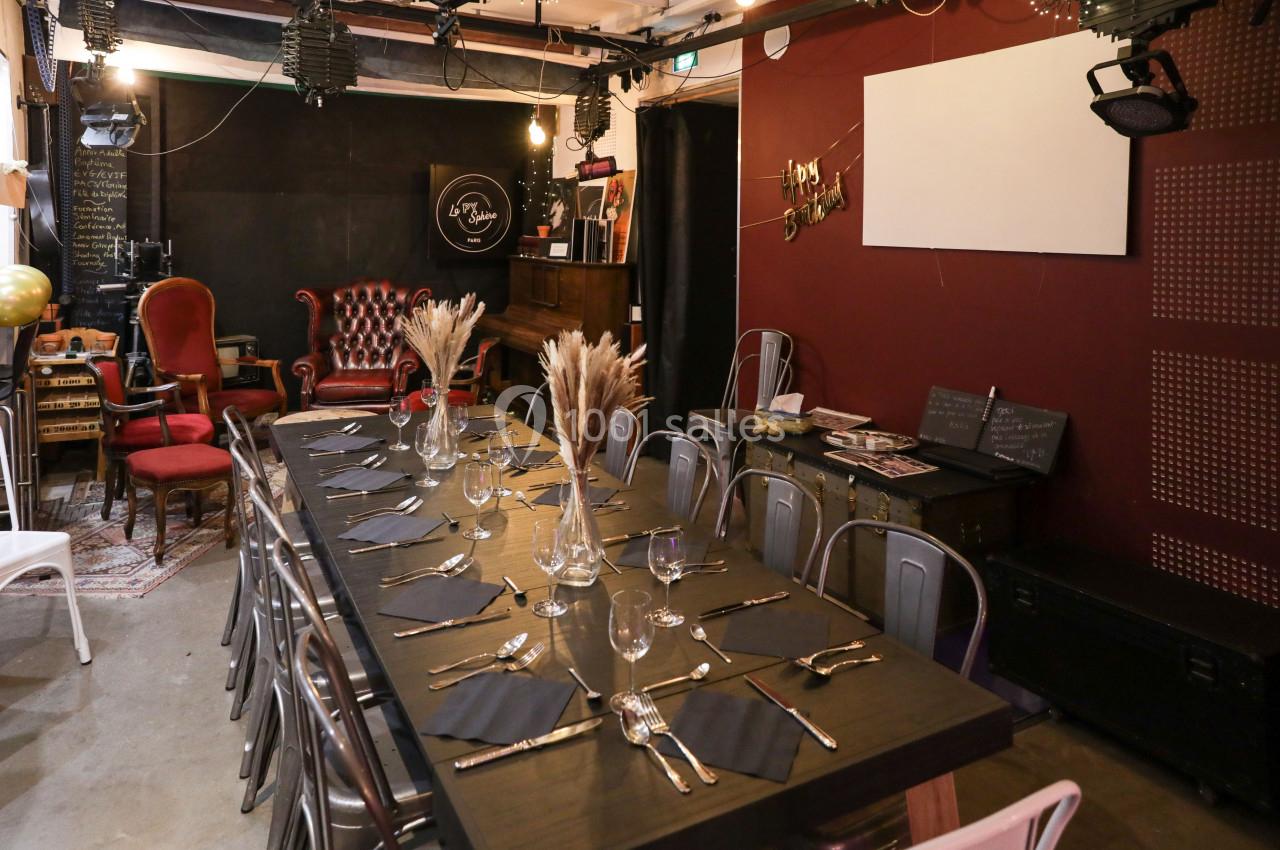 Salle décorée avec une grande table dressée, des chaises variées, et un coin salon avec fauteuils en arrière-plan.