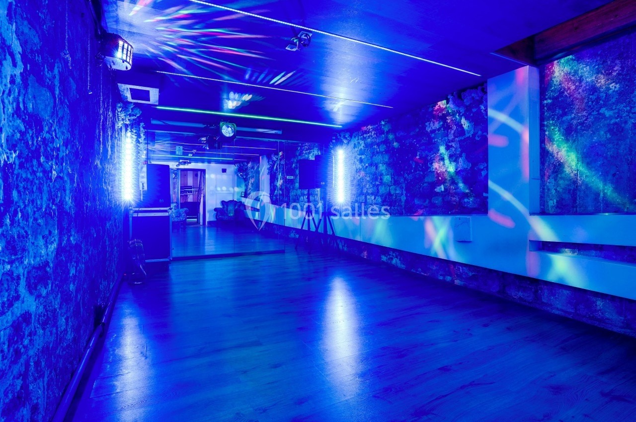 Salle avec éclairage bleu, murs en pierre, sol en bois et jeux de lumière colorés créant une ambiance festive.