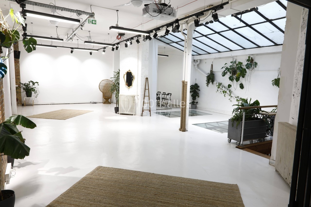 Espace lumineux avec sol blanc, verrière, plantes et mobilier minimaliste dans un décor moderne et épuré.