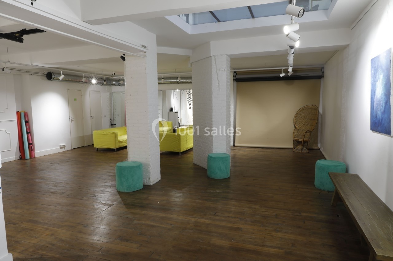 Espace intérieur lumineux avec parquet, canapés jaunes, poufs verts, et fauteuil en rotin près d'un mur beige.