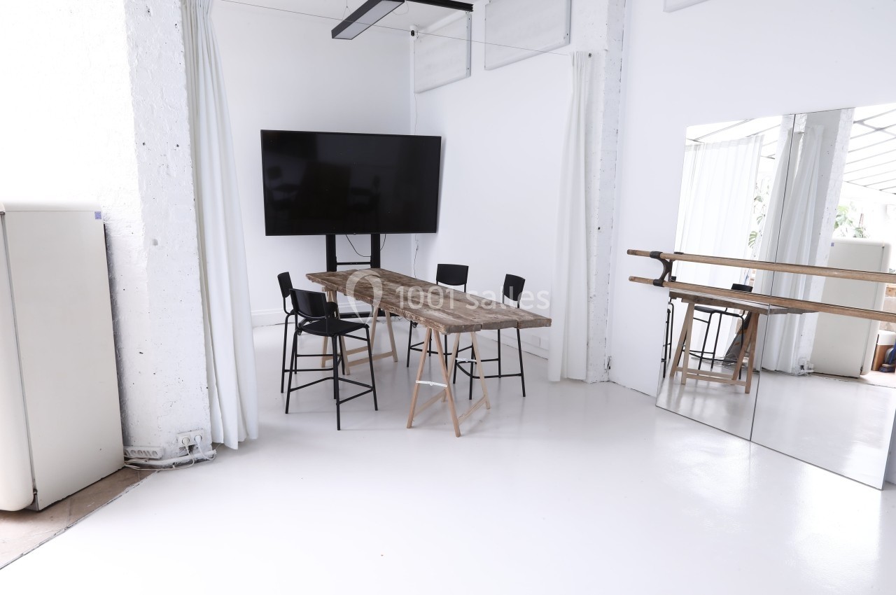 Salle lumineuse avec table en bois, chaises noires, écran mural et miroir sur le mur droit.