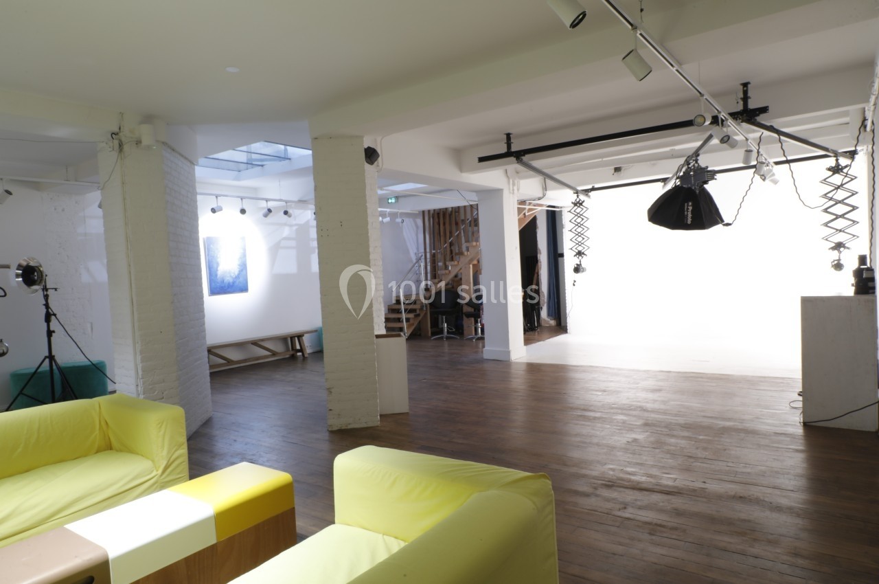 Studio photo lumineux avec fond blanc, équipements d'éclairage, parquet en bois et coin salon avec canapés jaunes.