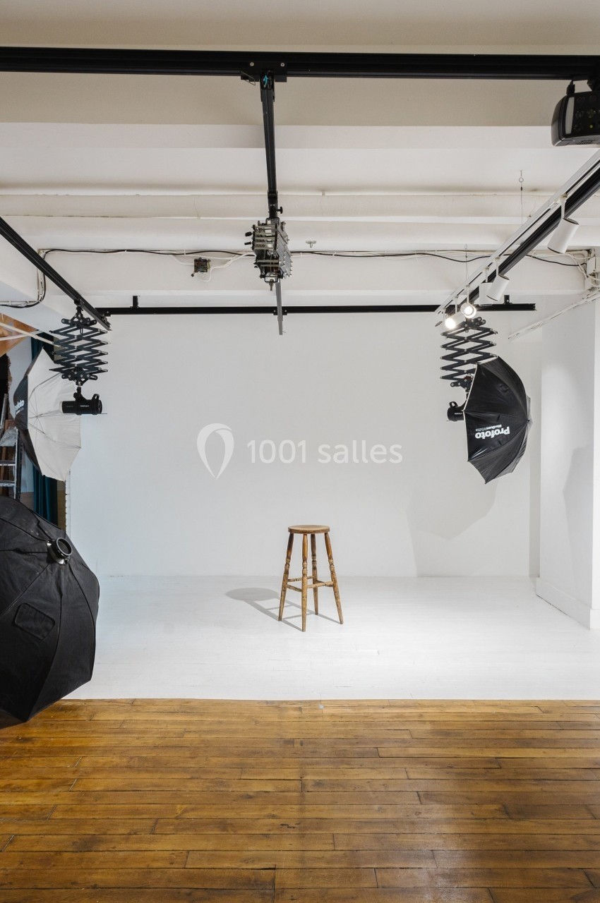 Studio photo avec fond blanc, tabouret en bois au centre, éclairages et parapluies noirs suspendus.