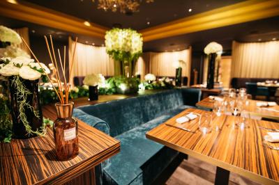 Terrasse en bois avec table haute en rotin, fleurs blanches et vue sur une salle de restaurant moderne éclairée.