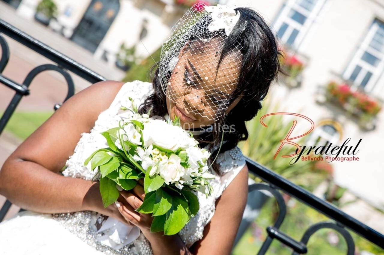 Une mariée tenant un bouquet de fleurs blanches, portant un voile en dentelle, devant un bâtiment en arrière-plan.