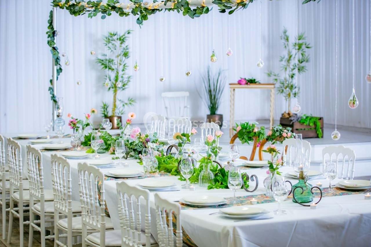 Table décorée pour un événement, ornée de plantes, fleurs et vaisselle élégante, avec fond blanc et végétation.