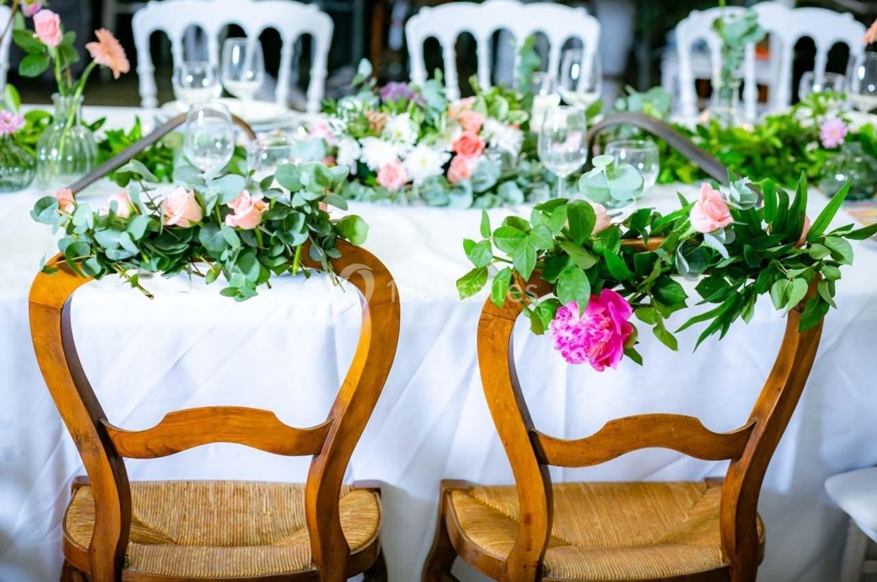 Chaises en bois décorées de feuillage et de fleurs roses, placées devant une table ornée de compositions florales.