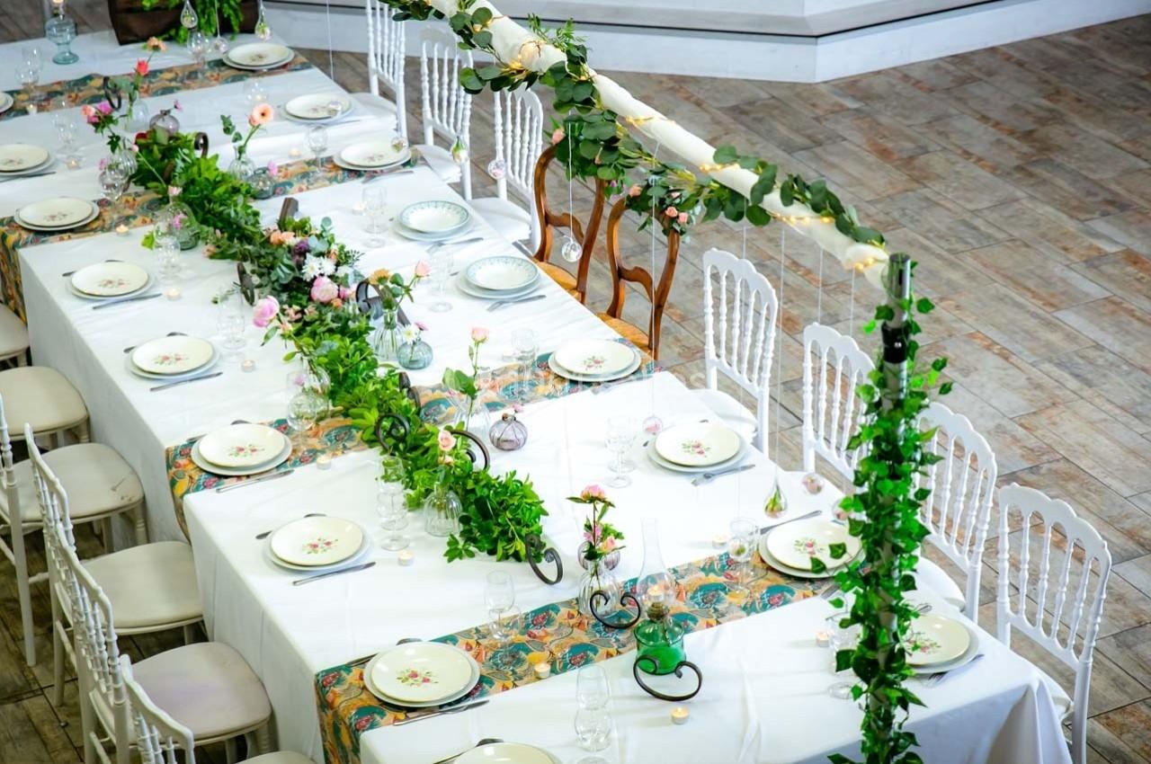 Grande table de réception décorée de nappes blanches, guirlandes végétales, vaisselle et fleurs colorées.