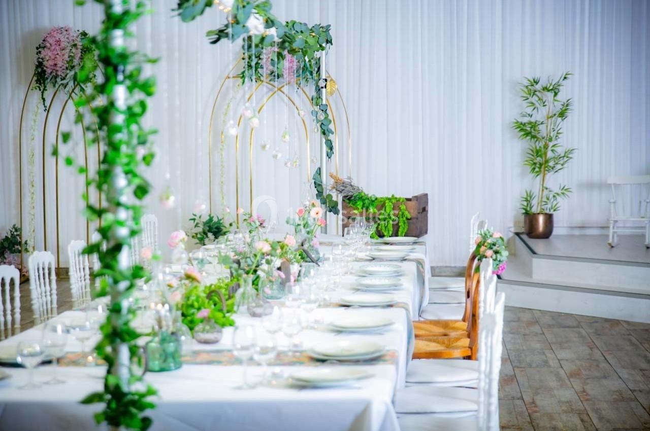 Table décorée avec vaisselle élégante, fleurs et feuillage, dans une salle lumineuse avec arches dorées et plantes.
