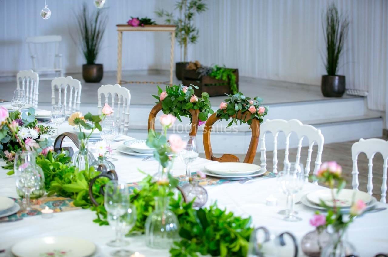 Table décorée avec des fleurs et de la verdure, entourée de chaises blanches dans une salle lumineuse et élégante.